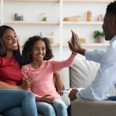 Positive black mother and daughter teenager at the appointment with male psychologist, happy african american girl giving psychotherapist high five, clinic interior, family therapy session concept